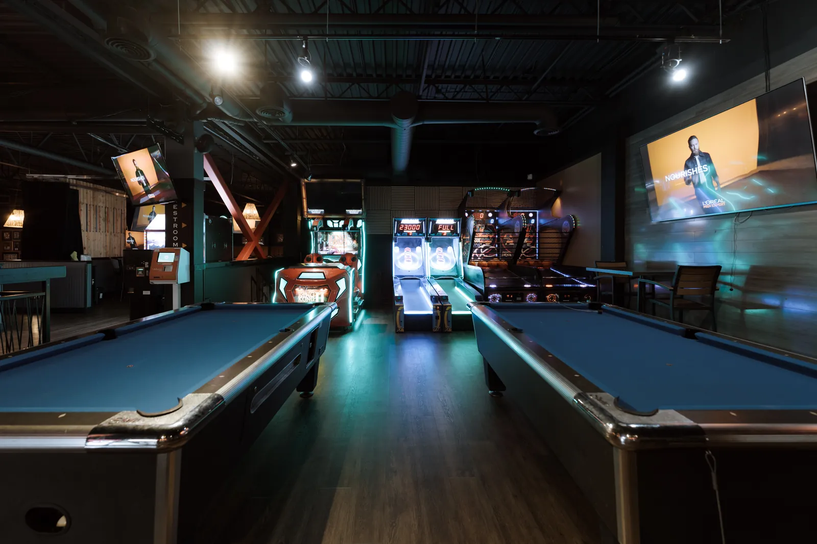 Two pool tables with blue felt, skee-ball lanes and arcade cabinets glowing behind them